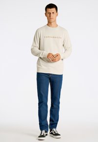 Sudadera de color beige claro con 'COPENHAGEN' impreso en beige oscuro, combinada con jeans azules y zapatillas en blanco y negro. Textura suave, corte casual.