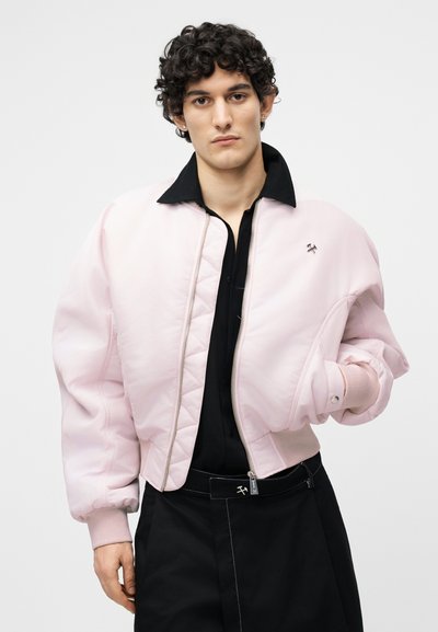 Νεαρός με σγουρά μαλλιά φοράει ένα ανοιχτό ροζ μπουφάν puffer πάνω από μια μαύρη πουκάμισο με γιακά και ψηλόμεσα μαύρα παντελόνια με λευκά ραφές.