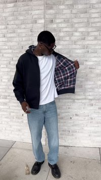 Hombre con gafas negras, sudadera negra con forro de cuadros, camiseta blanca, jeans azul claros y zapatos negros se encuentra de pie contra una pared de ladrillo blanco.