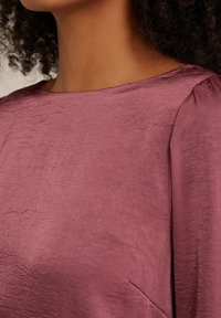 Krátký rukáv top z texturované, světle burgundské látky s kulatým výstřihem a jemným leskem. Obsahuje detail postranní kapsy.