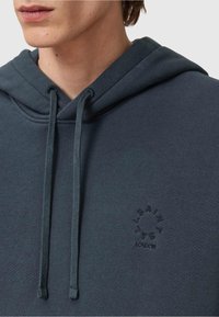 Tmavě šedá mikina s kapucí z texturované tkaniny, s centrálním logem na přední straně v kruhovém designu, se dvěma šňůrkami na stažení a uvolněným střihem.