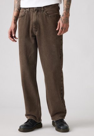 Brown denim jeans featuring a relaxed fit, five-pocket design, and visible stitching. Paired with black shoes and situated against a neutral background.