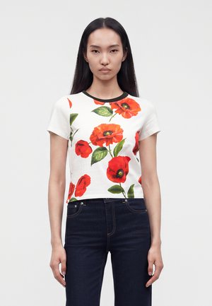 Femme aux longs cheveux noirs portant un t-shirt blanc à manches courtes avec de grandes fleurs de coquelicot rouges et un jean foncé, debout devant un fond uni.