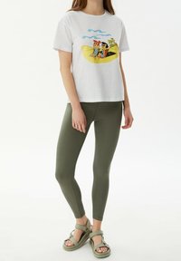Camiseta blanca de algodón con un estampado gráfico de dos personajes sobre un fondo amarillo; combinada con leggings verde oliva y sandalias beige.