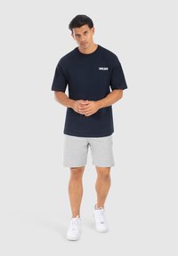 Camiseta de algodón azul marino de manga corta con el logo blanco "SMILODO", combinada con pantalones cortos grises claros y zapatillas blancas. Fondo neutro.