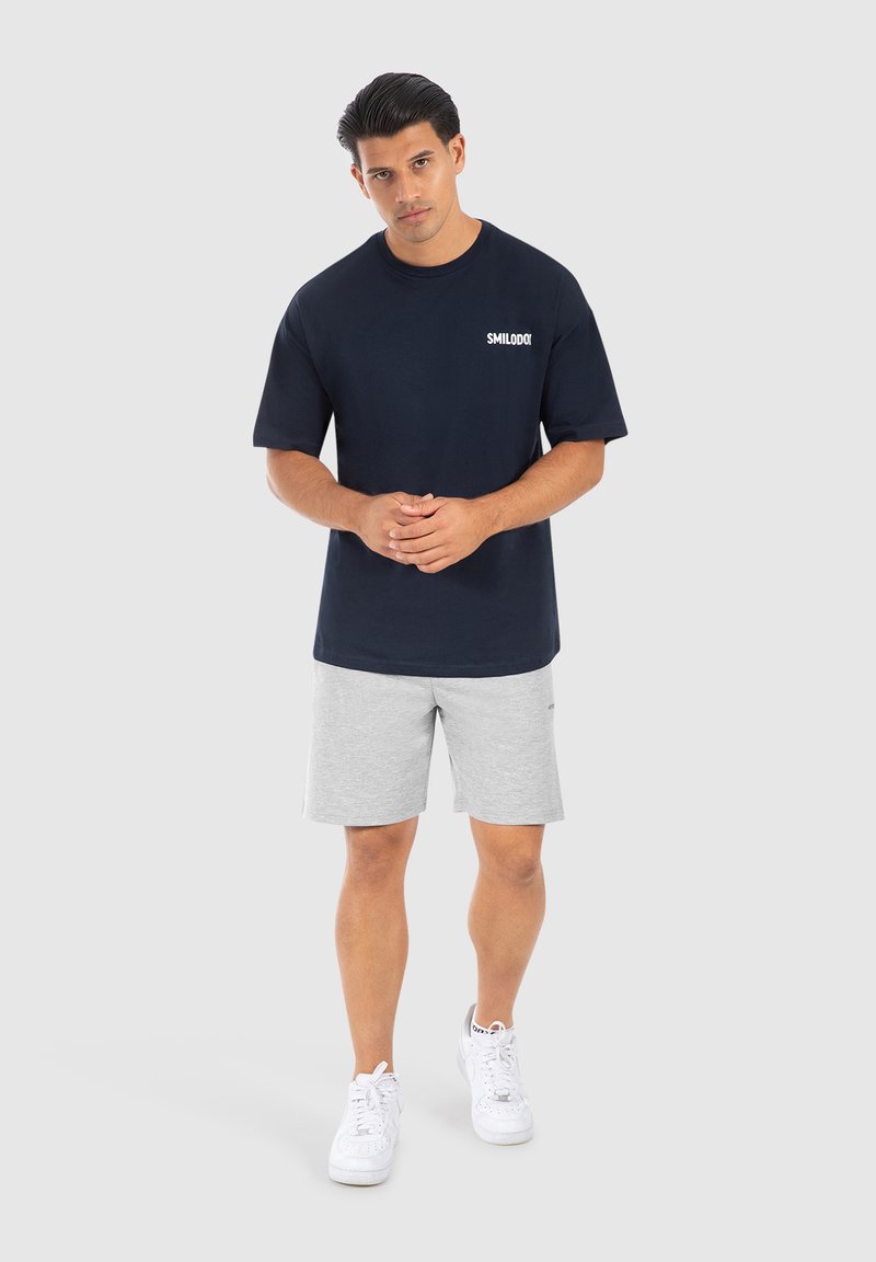 Camiseta de algodón azul marino de manga corta con el logo blanco "SMILODO", combinada con pantalones cortos grises claros y zapatillas blancas. Fondo neutro.