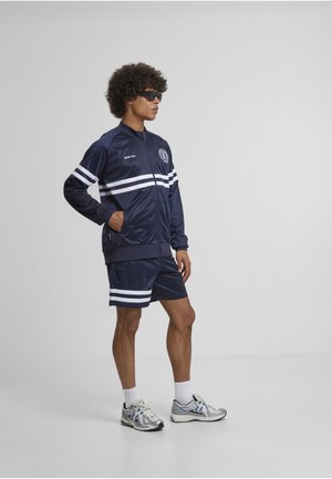 Man stands with hands in pockets wearing navy tracksuit jacket and shorts with white stripes, white socks, silver sneakers, and black sunglasses.