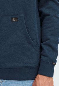 Sudadera azul hecha de una suave mezcla de algodón, con un bolsillo frontal y un dobladillo acanalado. Incluye un parche con el logo en el bolsillo y la manga.