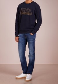 Granatowy sweter z logo "KARL LAGERFELD" w kolorze złotym, w zestawieniu z niebieskimi dżinsami i białymi trampkami z brązowymi podeszwami. Casualowy krój, bez widocznych wzorów.