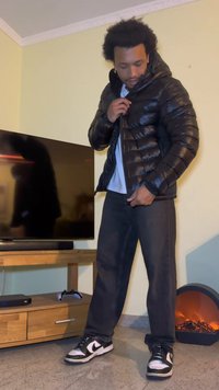 Črna puhovka s sijočim zaključkom, z zadrgo in visokim ovratnikom, nošena čez sivo srajco in temne kavbojke. Črno-bele superge.