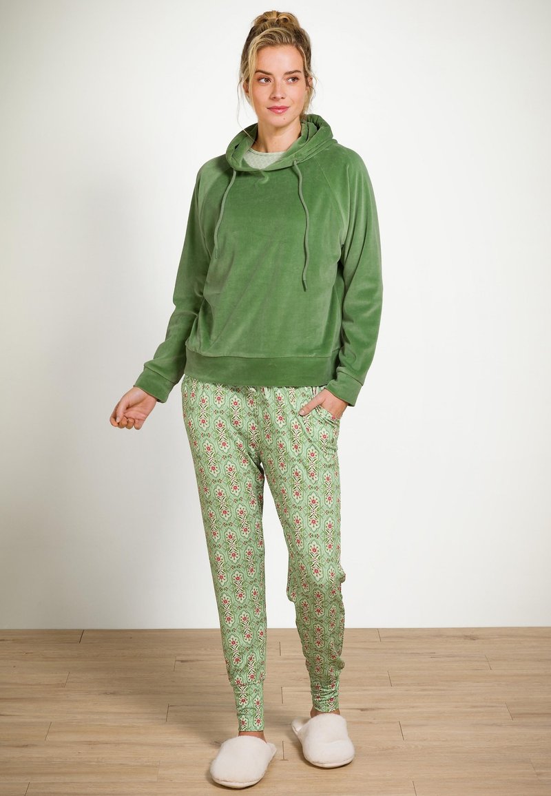Vrouw die binnen staat in een groene velour hoodie, met gemêleerde groene pyjamabroeken en witte slippers op een houten vloer.