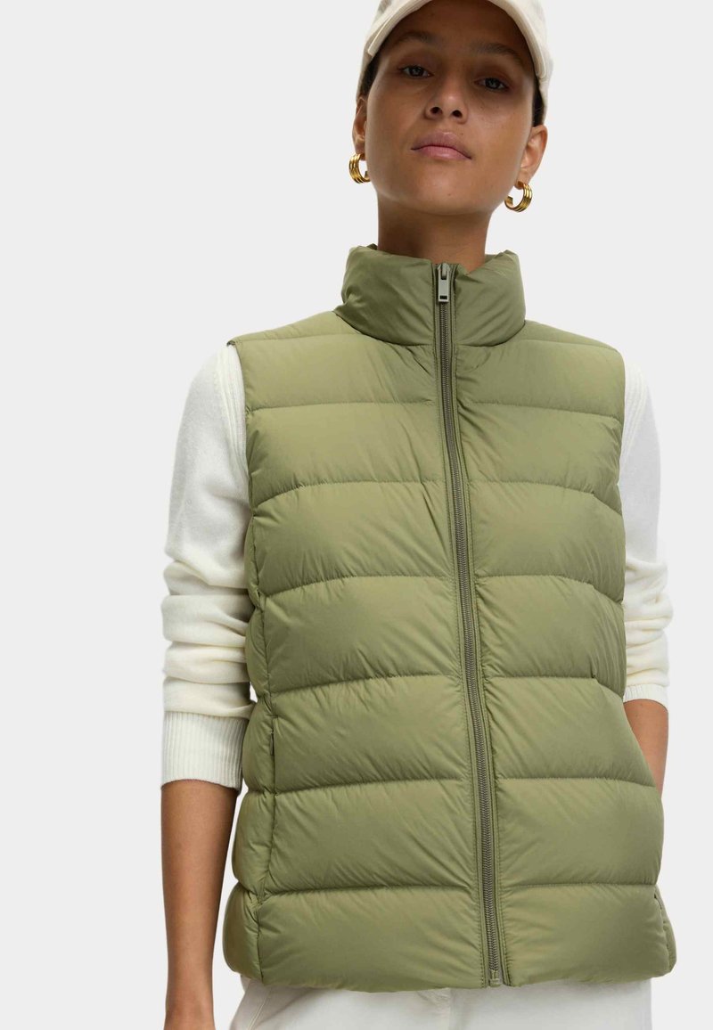 Groene puffer vest met horizontale quilting, hoge kraag en rits aan de voorkant. Draagt over een witte lange mouw top, heeft een gladde textuur.