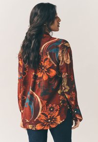 Long-sleeve shirt in rich burgundy with floral print in orange, blue, and green. Features a curved hem and button accents on cuffs.