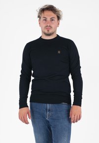 Maglione nero lavorato a maglia con scollatura rotonda, maniche lunghe e polsini a costine. Presenta un piccolo logo ricamato sul petto. Indossato con jeans blu.