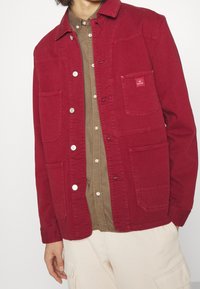 Veste en coton rouge avec un col classique, fermeture à boutons sur le devant, deux poches poitrine, et une chemise marron contrastante en dessous. Texture douce, design décontracté.