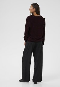 Donna con lunghi capelli scuri, che indossa un top a maniche lunghe bordeaux con motivo e pantaloni neri a gamba larga, in piedi e rivolta di lato su uno sfondo bianco.