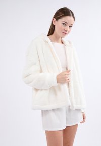 Biała kurtka z sztucznego futra z kapturem, charakteryzująca się miękką teksturą i zapięciem na zamek. Stylizowana z jasnymi szortami i podstawowym topem.