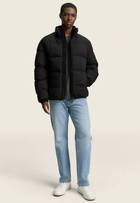 Črna puhaste jakna z visokim ovratnikom in sprednjim zadrgo. Kombinirana z svetlo modrimi kavbojkami in belimi supergami na nevtralni podlagi.