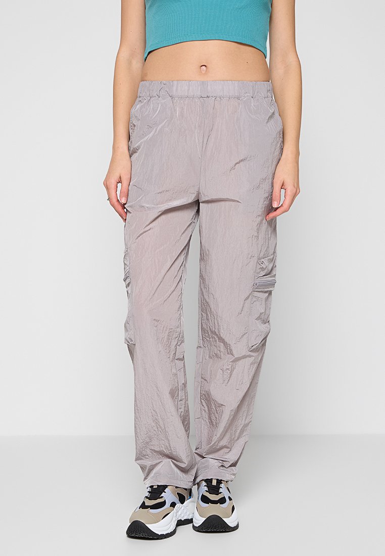 Femme portant un pantalon cargo gris clair froissé, un top court bleu sarcelle et des baskets épaisses blanches, beiges et noires, debout devant un fond uni.