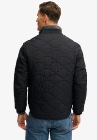 Veste matelassée noire rembourrée avec un motif de coutures ondulées, col montant et poignets côtelés. Portée avec un jean bleu, vue de dos.