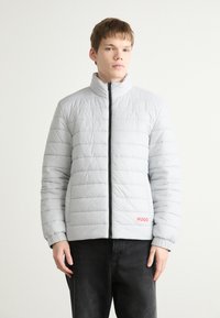 Gai pelēks pūkains mētelis ar augstu apkakli, vertikāli izšūtu dizainu, priekšējo rāvējslēdzēju un sarkano "HUGO" logo apakšējā kreisajā stūrī.