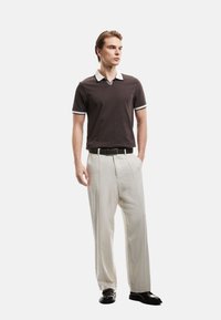 Bruine polo met een witte kraag en manchetten, gecombineerd met lichtbeige broek. Een zwarte riem en schoenen maken de outfit compleet. Eenvoudige ontwerpelementen.