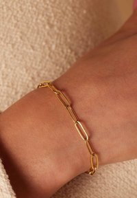Gouden kettingarmband met langwerpige, verbonden ovale vormen, rustend op een huidkleurige pols. Zachte, gestructureerde achtergrond.