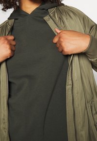 Kurtka bomberka w oliwkowej zieleni z zamkiem z przodu, noszona na ciemnozielonym kapturze. Kurtka posiada ściągacze przy mankietach i gładką teksturę.