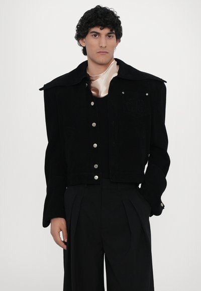 Black corduroy cropped jacket with large collar, silver hardware, and front pockets. Worn over a black outfit with a satin, light-colored neck tie.