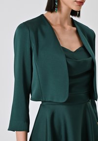 Giacca di raso teal a tre quarti, con colletto strutturato e texture liscia, indossata sopra un top a collo drappeggiato coordinato.