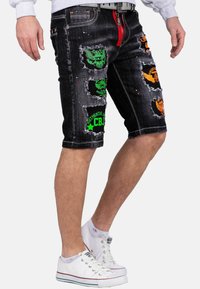 Shorts in denim neri con dettagli sdruciti, caratterizzati da toppe colorate in verde, arancione e giallo. Include una cerniera rossa e cuciture bianche.