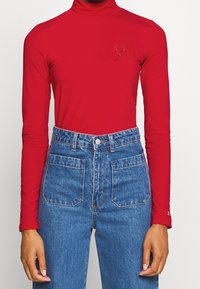 Red long-sleeve turtleneck top with a fitted design and subtle embroidered detail on the left chest, paired with high-waisted blue jeans.