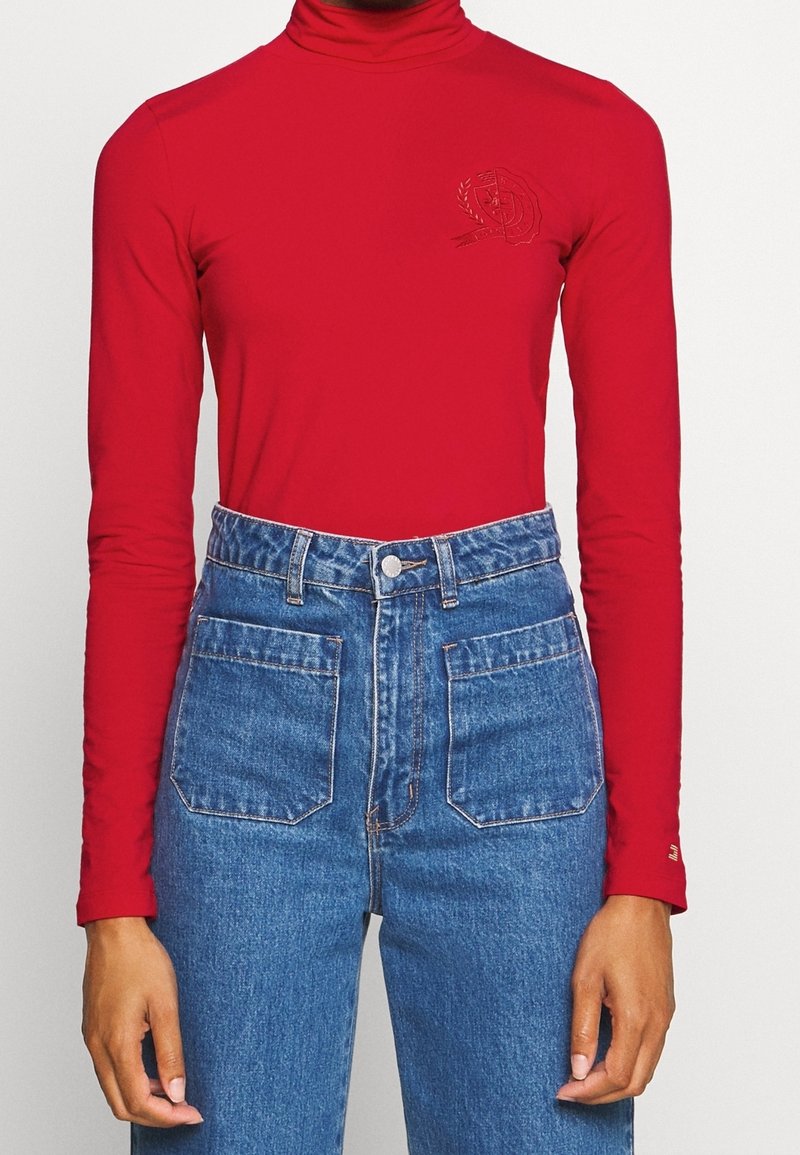 Red long-sleeve turtleneck top with a fitted design and subtle embroidered detail on the left chest, paired with high-waisted blue jeans.