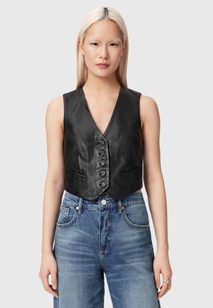 Mujer con cabello rubio largo que lleva un chaleco negro de cuero con botones y jeans de mezclilla azul contra un fondo liso gris claro.