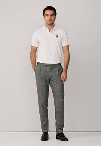 Vit pikétröja med en liten logotyp, kombinerad med grå rutiga byxor. Modellen står i svarta skor och visar upp en skräddarsydd passform.