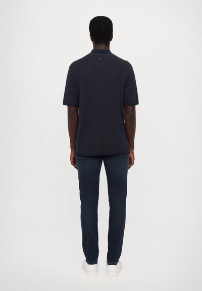 Marineblå, ribbestrikket kortermet polo skjorte med krage, kombinert med mørke jeans og hvite sneakers, sett fra baksiden.
