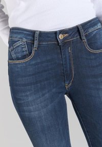 Unos vaqueros de mezclilla azul oscuro con un ajuste slim, que cuentan con costuras doradas, un bolsillo frontal y un cierre de botón en la cintura.