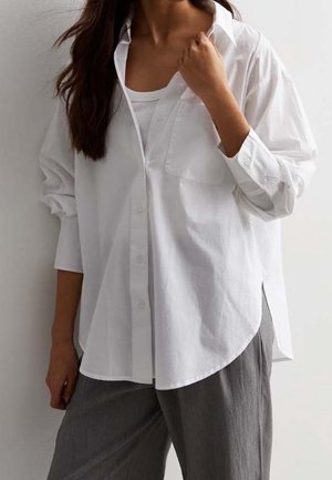 Kobieta ubrana w luźną białą koszulę na guziki na białym tank topie i szarych spodniach, ręką poprawiająca kołnierz koszuli.