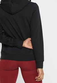 Felpa nera con cappuccio, maniche lunghe, polsini a coste e orlo dritto. Il tessuto appare morbido; indossata con pantaloni bordeaux.