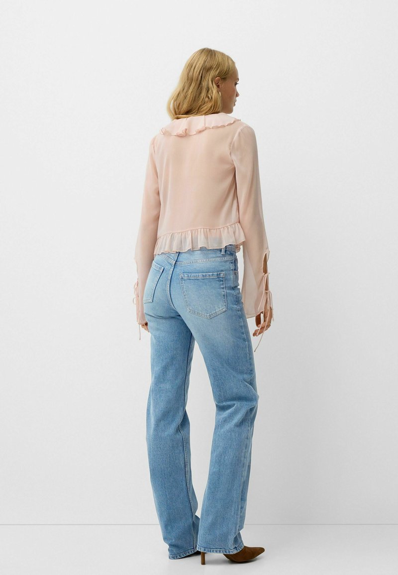 Helles rosa, durchscheinendes Blusenshirt mit Rüschenkragen und -saum, langen Ärmeln mit gebundenen Akzenten, kombiniert mit hellblauen, weit geschnittenen Jeans und braunen Absätzen.