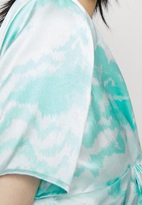 Jasnoniebieska bluzka z krótkim rękawem, ozdobiona wzorem tie-dye w odcieniach turkusu i bieli, z detalem w postaci wiązania w talii oraz gładką teksturą.