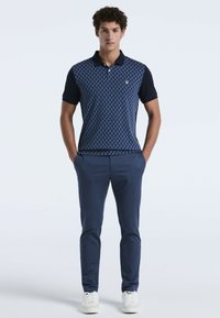 Marineblauw polo-shirt met geometrisch patroon aan de voorkant, korte mouwen en een kraag, gecombineerd met marineblauwe broeken en witte sneakers.