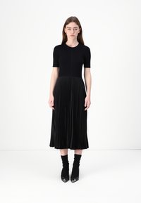 Abito midi nero con maniche corte, corpino aderente e gonna plissettata. Realizzato in tessuto morbido, presenta una trama a strati e scarpe lucide.