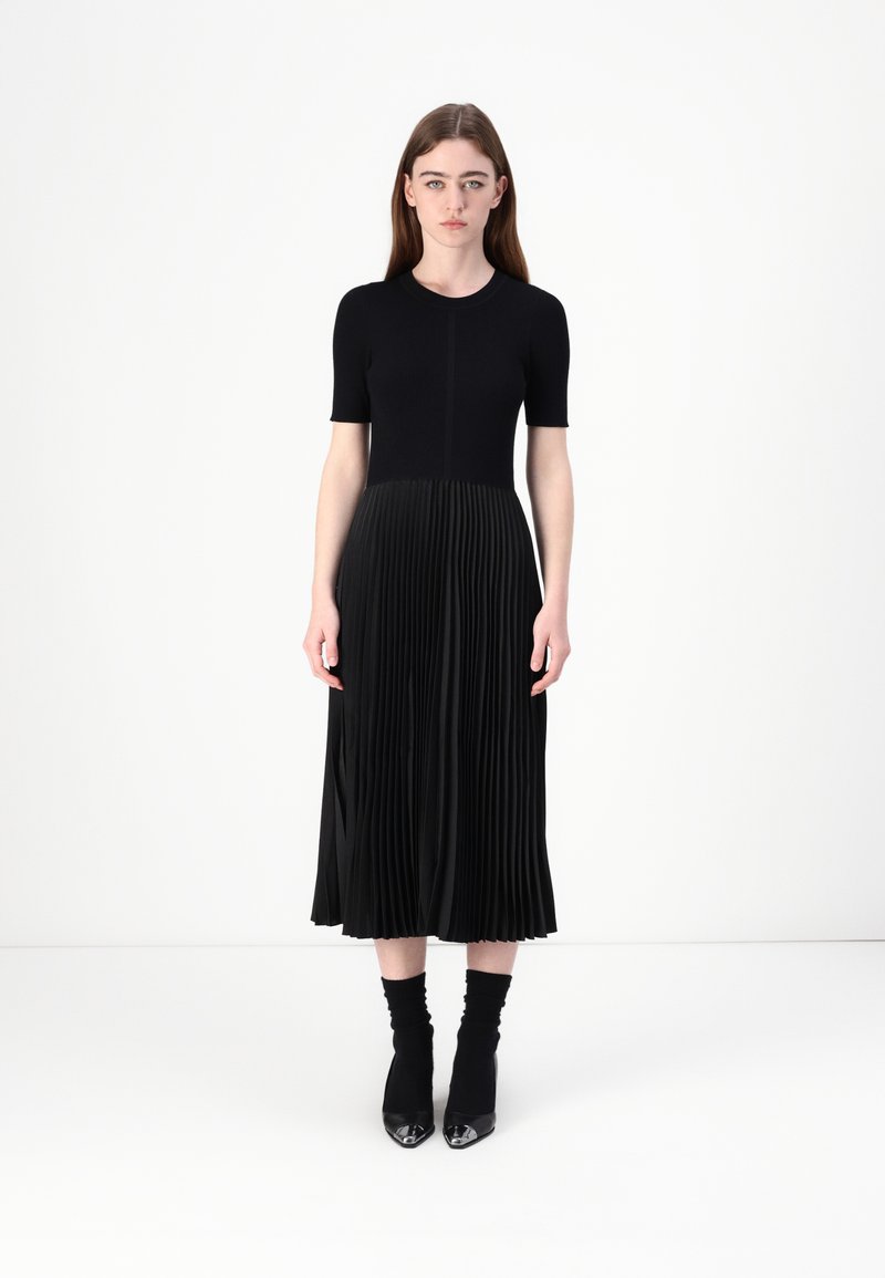 Abito midi nero con maniche corte, corpino aderente e gonna plissettata. Realizzato in tessuto morbido, presenta una trama a strati e scarpe lucide.