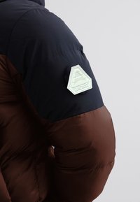 Chaqueta acolchada marrón y negra con textura acolchada y un parche de logo blanco hexagonal en el hombro izquierdo.