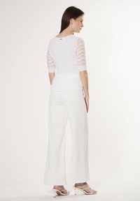 Weißes Rippenstrickoberteil mit durchsichtigen gestreiften Ärmeln, taillierter Silhouette und rundem Rücken. Kombiniert mit weitgeschnittenen weißen Hosen und minimalistischen Absatzsandalen.