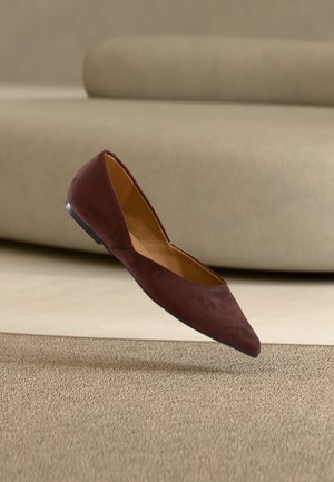 Mørkebrun semsket spiss tå flat sko som svever over beige teppe med en beige sofa i bakgrunnen.