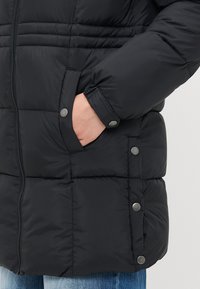 La mano di una persona nella tasca di un giubbotto puffer nero con bottoni a pressione sul lato e sul polsino, indossando dei jeans blu.