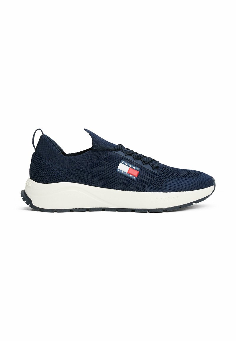 Baskets tricotées bleu marine avec semelle intermédiaire blanche et accents texturés. Comprend des lacets plats, une languette de tirage et un petit patch logo en rouge et bleu.