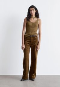 Brown ribbed tank top with buttons and brown corduroy flared pants, complemented by a green necklace and gray shoes. Simple design.